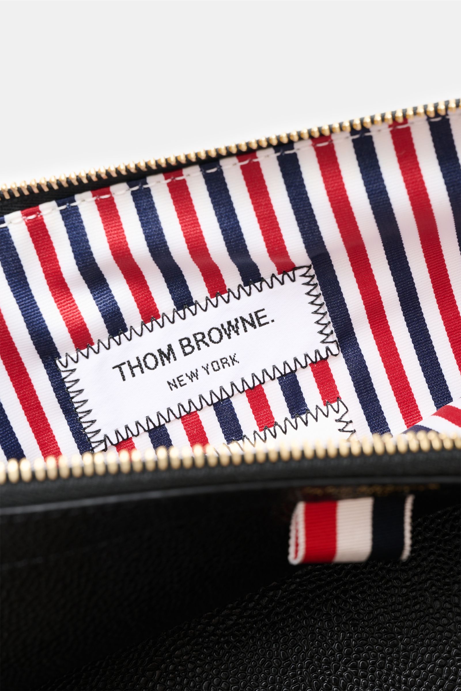 Nahaufnahme des Innenfutters der Thom Browne Dokumentenmappe schwarz mit Streifendesign und THOM BROWNE Label, goldfarbener Reißverschluss.