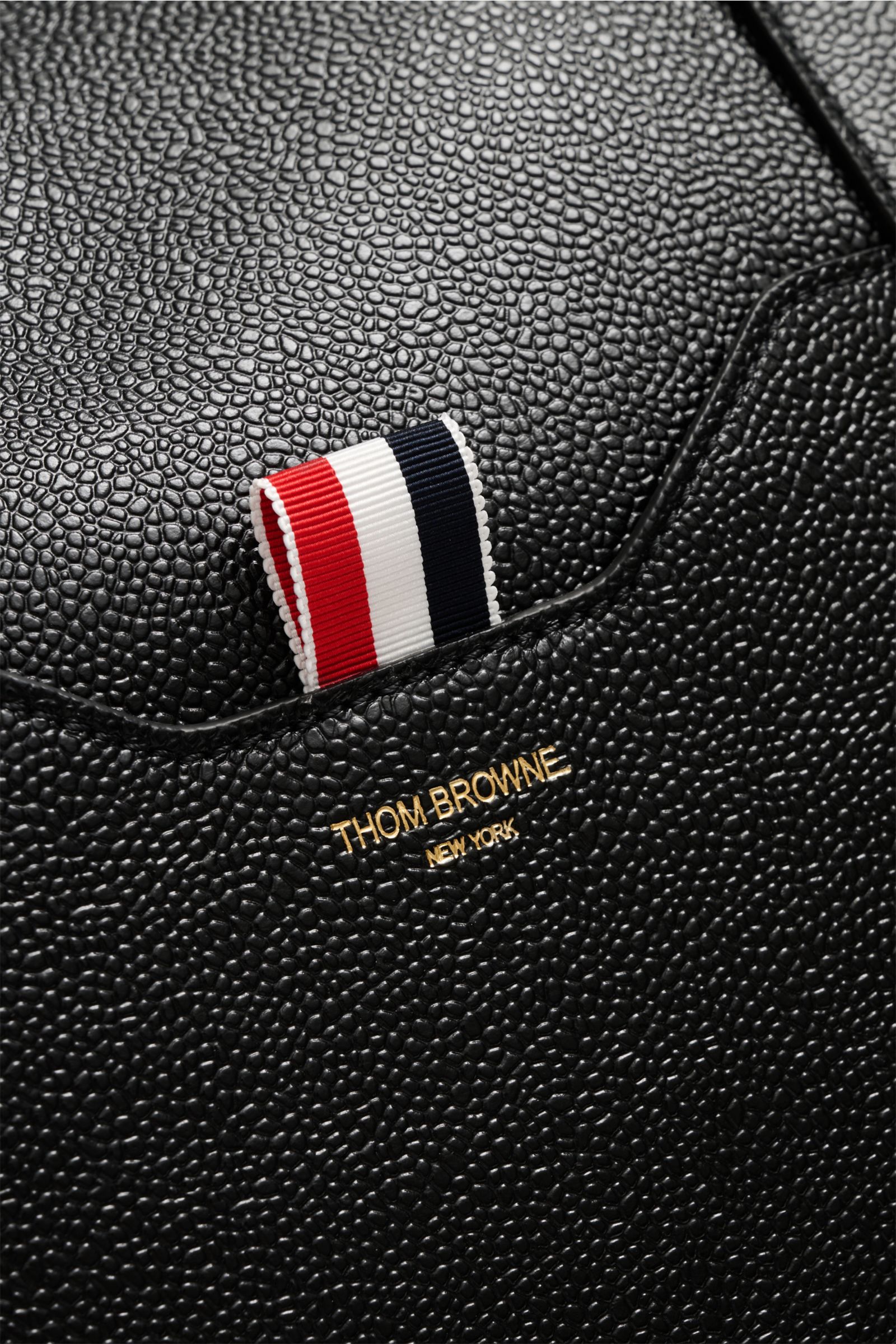 Nahaufnahme des genarbten Leders und der Logo-Prägung des Thom Browne Weekender schwarz, mit Detail des markanten roten, weißen und blauen Webbands.