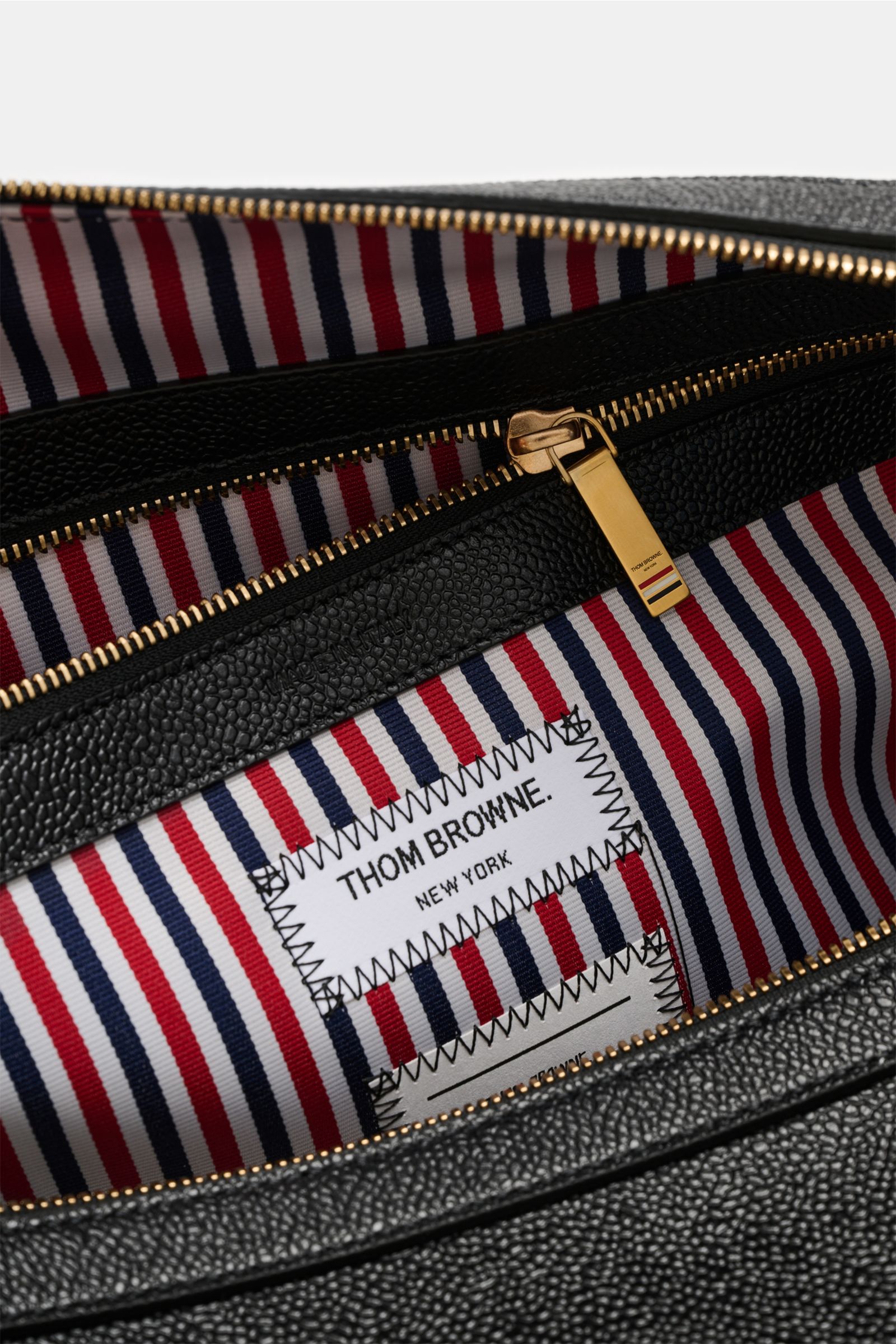 Nahaufnahme des geöffneten Thom Browne Weekender schwarz aus genarbtem Leder, zeigt goldfarbenen Reißverschluss und gestreiftes Innenfutter.
