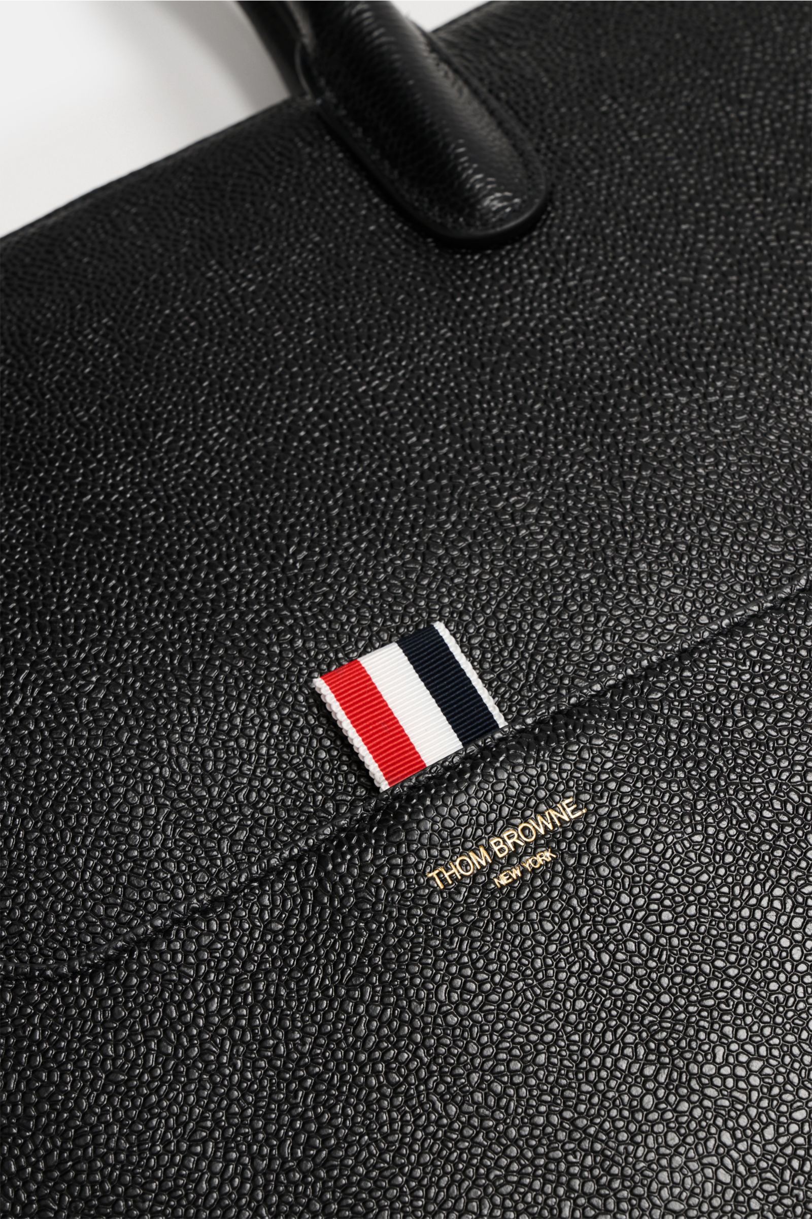 Nahaufnahme des fein genarbten Leders und robuster Tragegriffe des Thom Browne Shopper schwarz, mit goldfarbener Logo-Prägung und Tricolor-Detail von oben fotografiert.