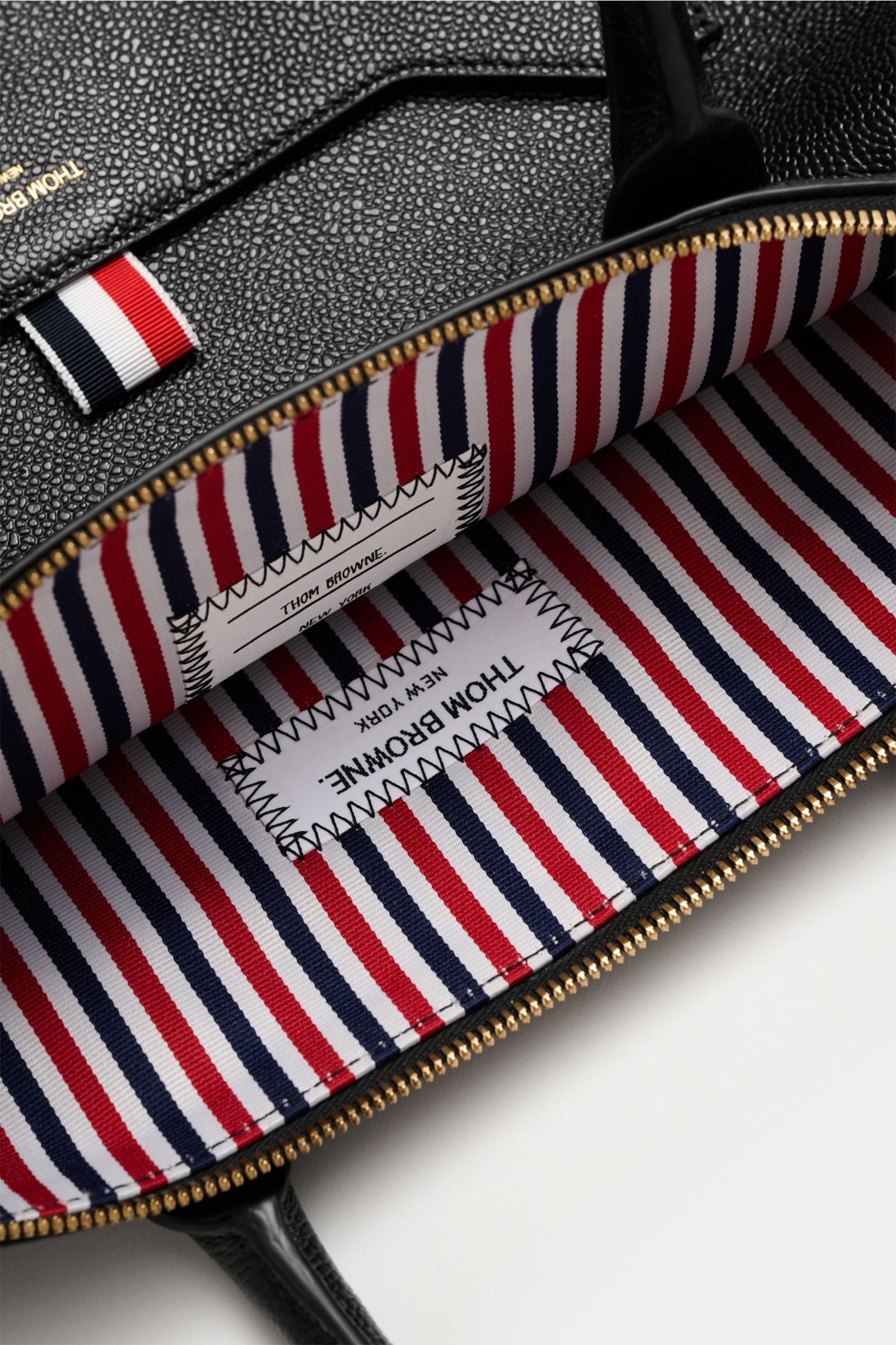Nahaufnahme des geöffneten Thom Browne Shopper schwarz aus genarbtem Leder, gezeigt von oben auf das gestreifte Innenfutter mit fünf Innenfächern, goldfarbener Reißverschluss, robusten Tragegriffen und regulierbarem Schultergurt.