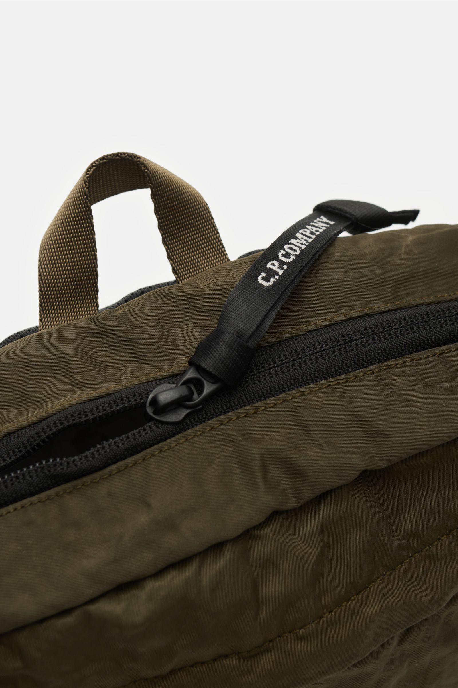 Close-up top view of C.P. Company Belt bag olive showing water repellent microfibre with subtle sheen, zip with branded pull, and robust carry loop. Don’t go on trips or into the city without this compact, piece-dyed belt bag made of water repellent mi