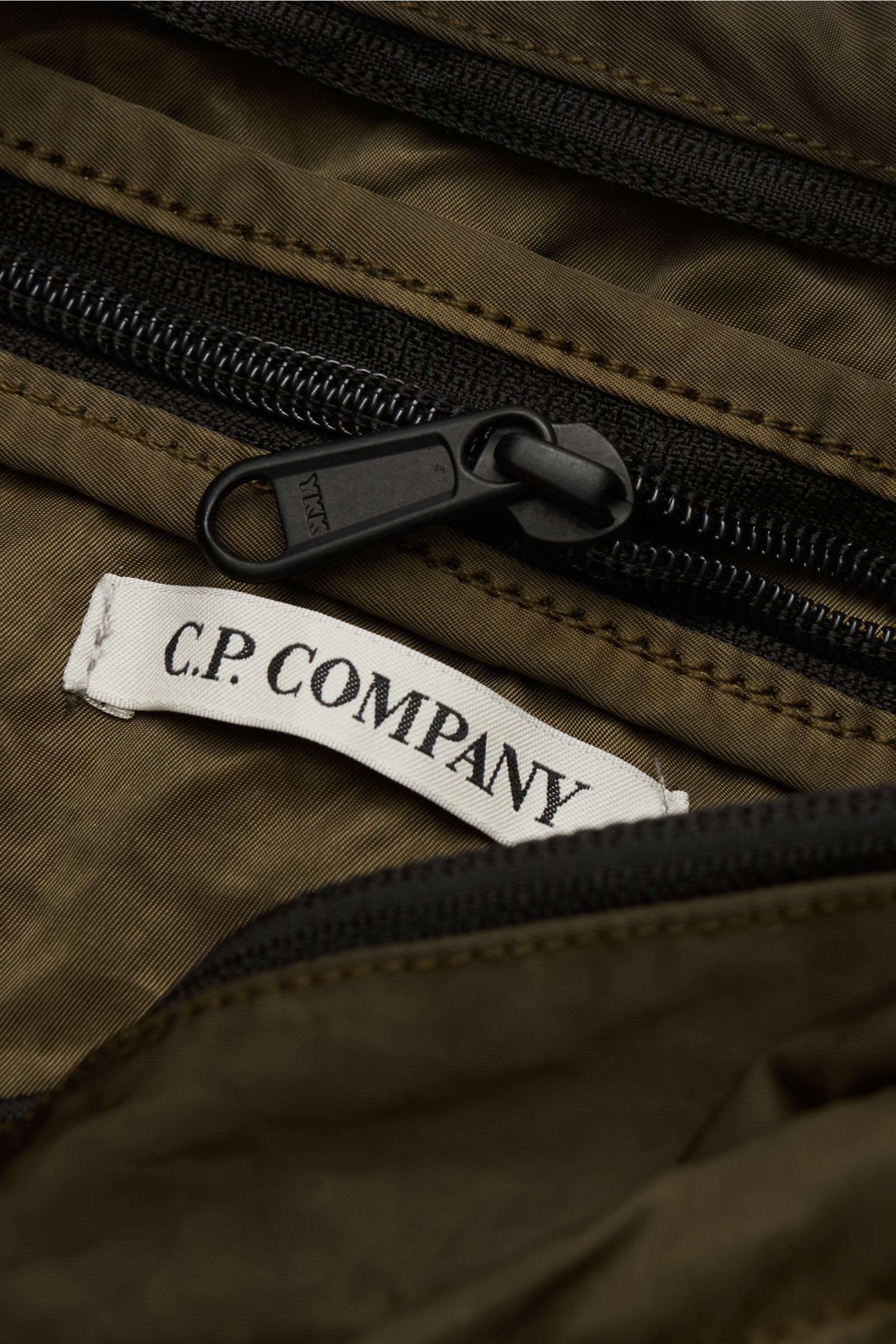 Close-up view of the C.P. Company Belt bag olive showing black zipper and white brand label on water repellent microfibre fabric. Don’t go on trips or into the city without this compact, piece-dyed belt bag made of water repellent microfibre by C.P. COMPA