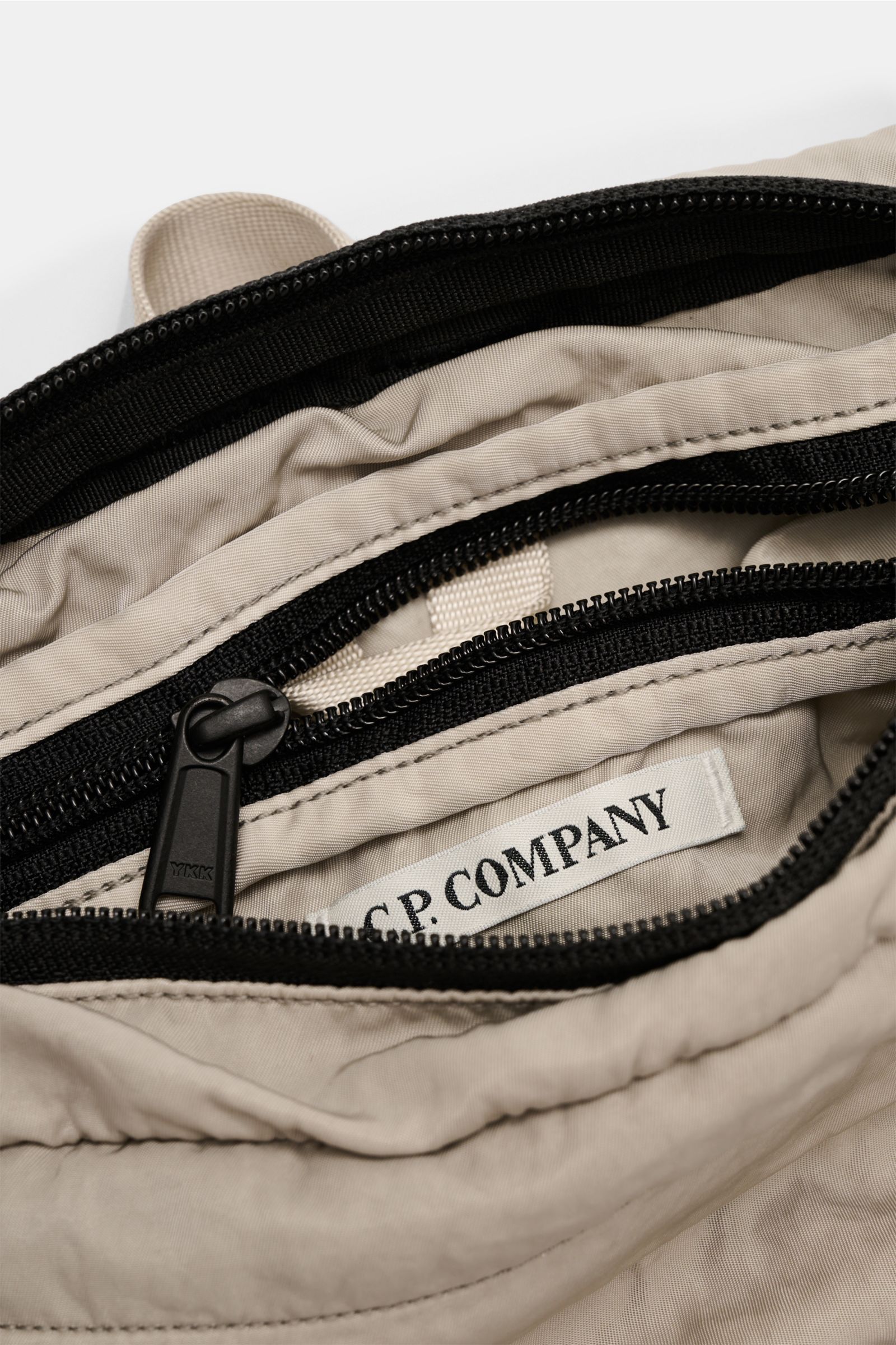 Nahaufnahme der geöffneten C.P. Company Gürteltasche grau mit wasserabweisender Microfaser, Reißverschlüssen und Innenfach, von oben fotografiert.