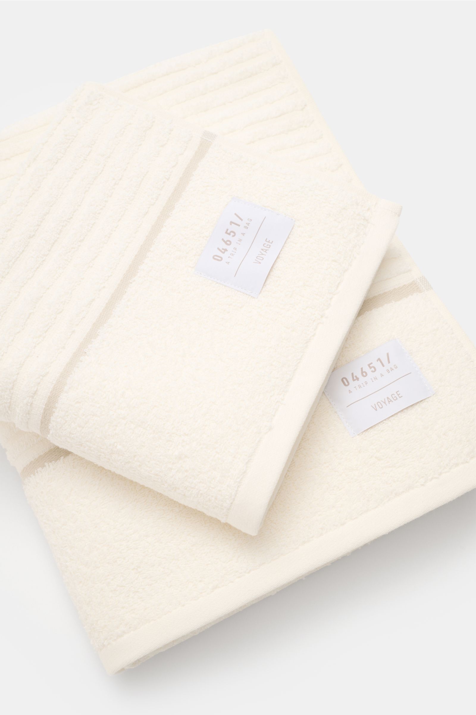 Close-up top-down view of two cream and white towels with '04651/ A trip in a bag VOYAGE' labels, part of the 04651/ A trip in a bag Reise-Set 'Voyage' creme/weiß.