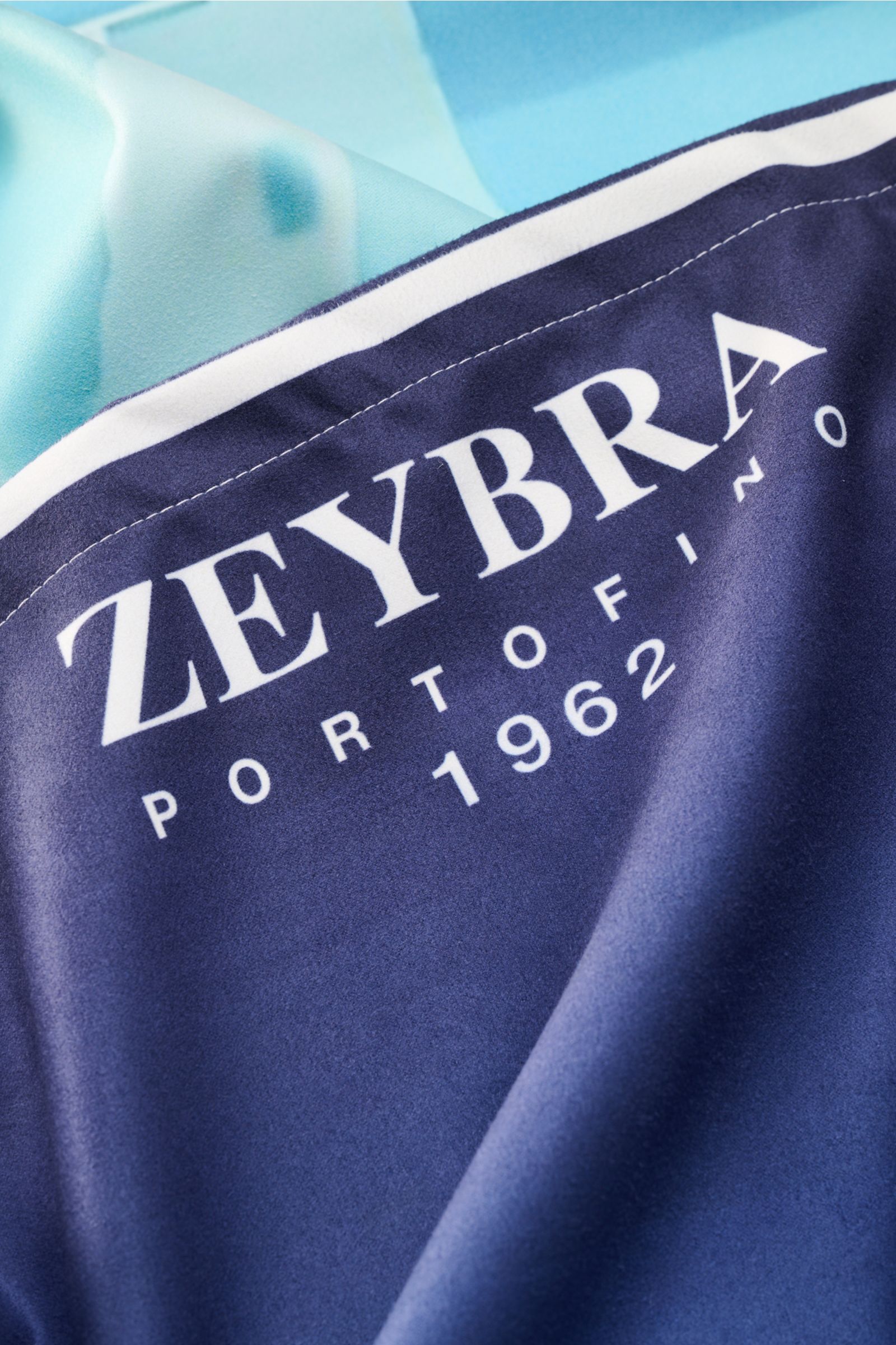 Nahaufnahme aus schräger Perspektive des Zeybra Portofino 1962 Strandtuch blau/multicolor gemustert mit weißem Schriftzug, feiner Microfaser, softem Griff, All-Over-Print mit Auto, ca. 120x190 cm.