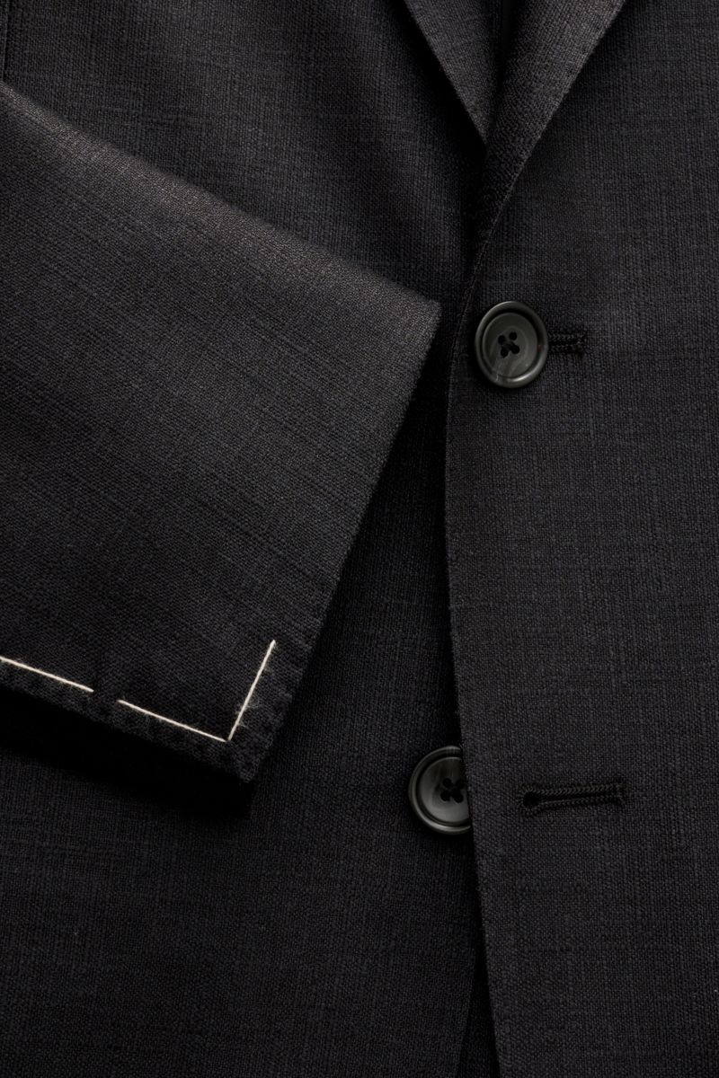 Close-up front view of the Tagliatore Smart-casual jacket 'Monte Carlo' black, showing slim fit, two-button style, step lapel, signature pin, breast pocket, patch hip pockets, unfinished sleeve cuffs, and fine textured silk and virgin wool blend fabric. 
