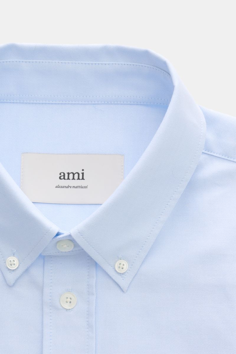 A close-up front view of an Ami Paris Oxford shirt button-down collar light blue, showing the button-down collar, patch button placket with mother-of-pearl buttons, and signature heart stitching. For relaxed casual styles and more formal looks: This elega