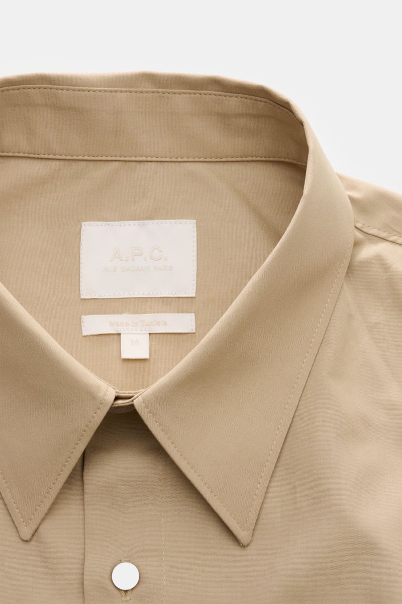 A close-up front view of the A.P.C. Kurzarmhemd Kent-Kragen sand, a sand-colored short-sleeve shirt with a Kent collar, made of pure cotton, featuring a smooth texture, oversized fit, French button placket, patch chest pocket, back yoke with pleat, and a 