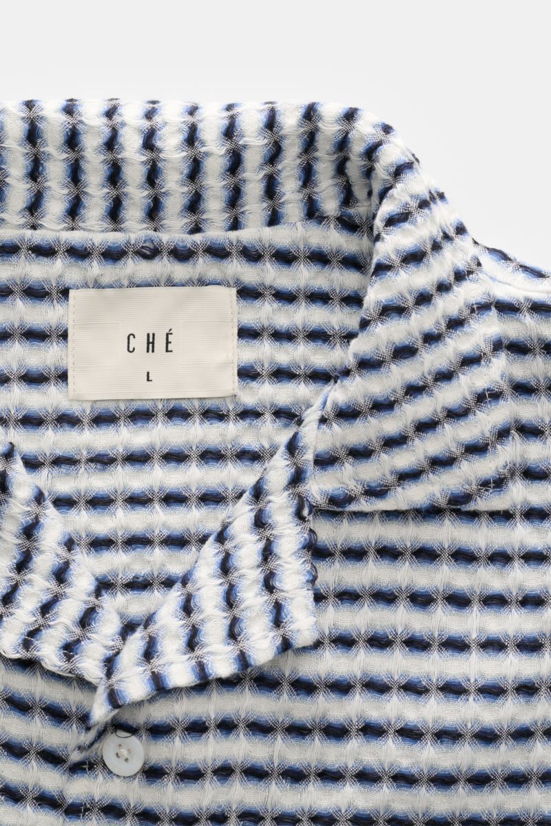 Close-up front view of Ché Leinen-Kurzarmhemd 'Riita' Kubanischer Kragen blau/weiß gemustert showcasing its intricate blue and white woven waffle piqué pattern and the CHÉ label. 

Description: Im Sommer kreiert das Leinen-Kurzarmhemd 'Riita' mit Kuban