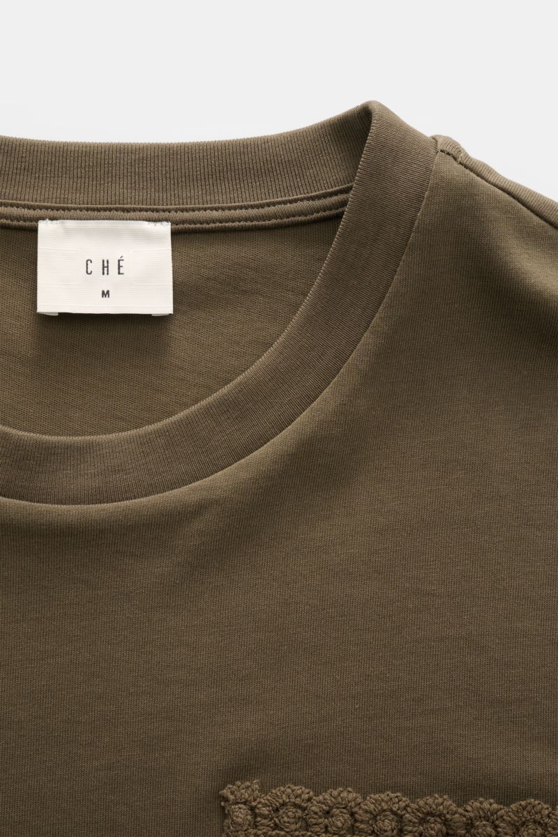 Close-up front top view of Ché T-Shirt 'Inigo' dark olive, showing round neckline, soft cotton fabric, and crochet detail at hem. Sommerliche Lässigkeit: Die Das 'Inigo' T-Shirt von CHÉ wird mit seinen Häkel-Details zum stilvollen Blickfang für entspannte
