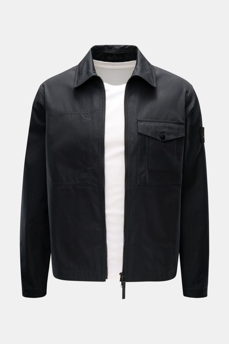 Front view of Stone Island Overshirt 'Ghost Piece' black, a slim-fit black cotton canvas jacket with turn-down collar, two-way zip, breast pocket with flap, tonal sleeve badge, and button cuffs, worn over a white shirt. Function meets comfort: This STONE 