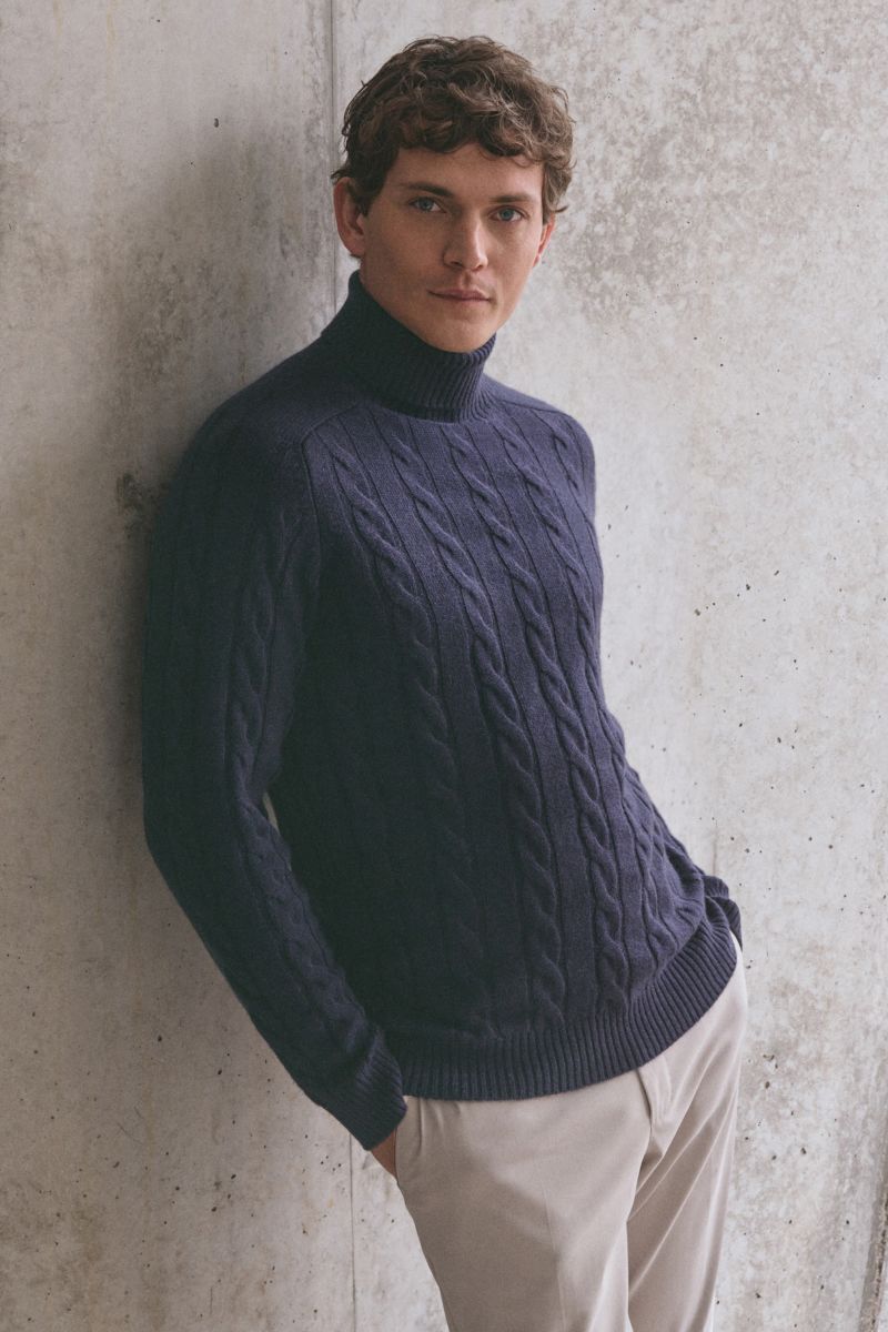 Mann lehnt seitlich an Wand, trägt von Braun Cashmere Rollkragenpullover navy mit Zopfstrickmuster, Regular Fit, weicher Griff, Rollkragen.