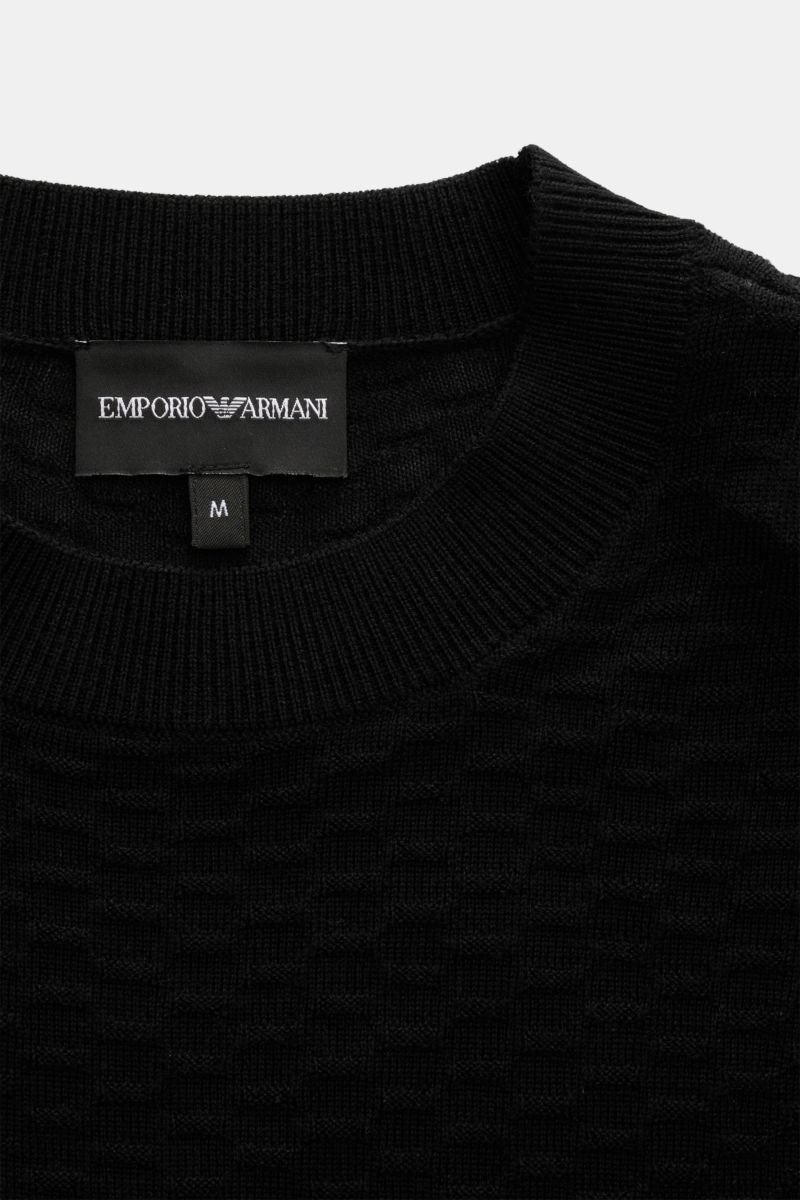 Close-up front view of the Emporio Armani Rundhalspullover schwarz, a lightweight black crewneck sweater made of pure new wool with a subtle textured knit pattern, soft touch, regular fit, ribbed collar and cuffs, and a woven label at the hem. Vielseitige