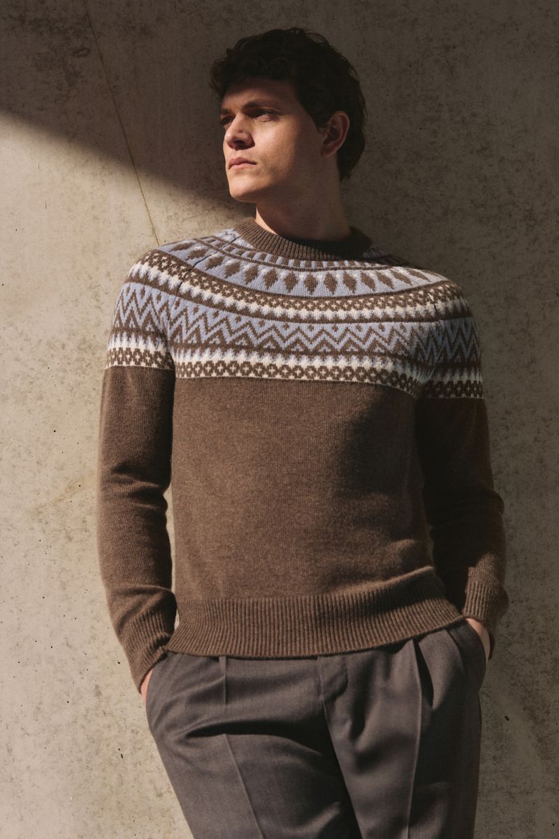 Mann lehnt seitlich vor Betonwand, trägt von Braun Cashmere Rundhalspullover braun/rauchblau/creme gemustert mit Norweger-Strickmuster, Slim Fit, Rundhals und Raglanärmel.