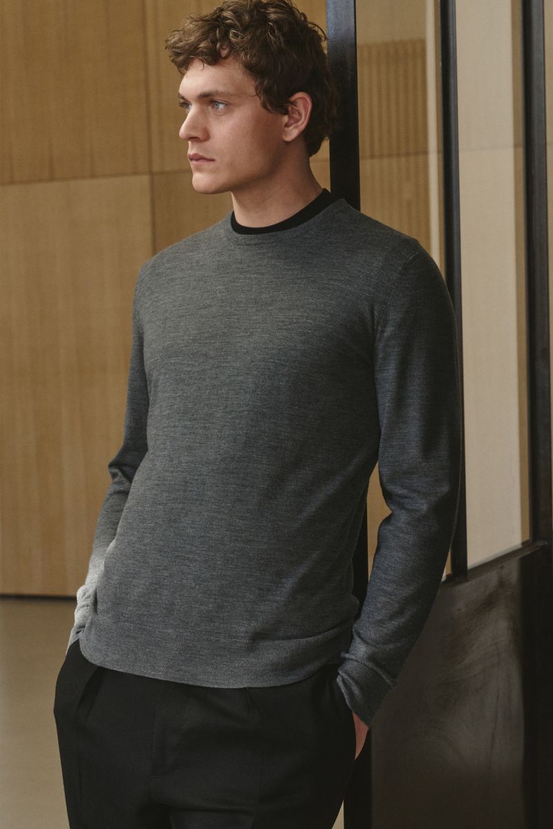 von Braun Merino Rundhalspullover anthrazit meliert, Slim Fit, aus reiner Merinowolle, feines Strickbild, Rundhalsausschnitt, elastische Rippbündchen, frontal seitlich fotografiert, Modell lehnt an Wand