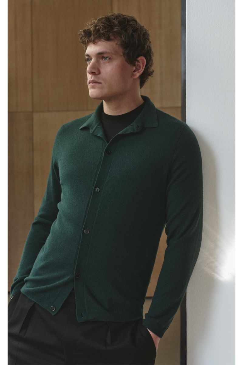 Mann von schräg vorne lehnt an Wand, trägt von Braun Cashmere Strickhemd dunkelgrün, Slim Fit, reiner Cashmere, glatte Knopfleiste, klassischer Kragen, Rippbündchen.