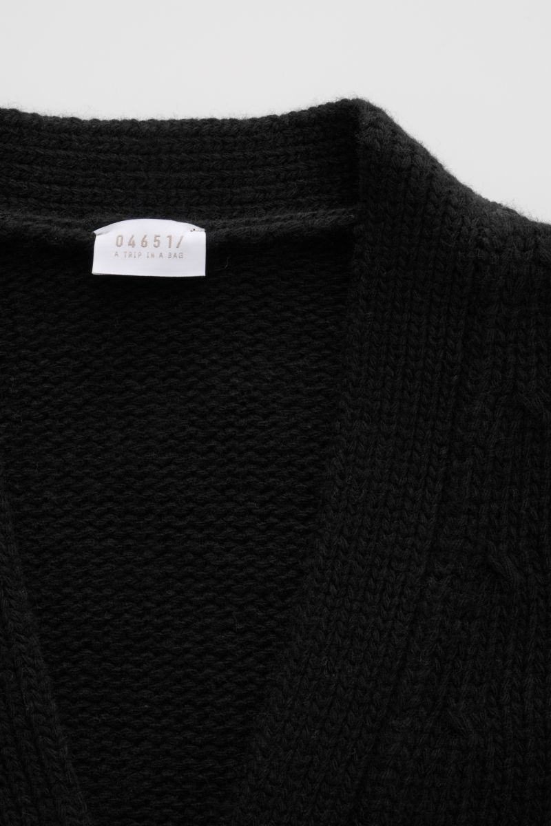 Nahaufnahme des schwarzen 04651/ A trip in a bag Cardigan 'Bold' mit grobem Strickbild, weichem Griff und sichtbarem Label innen am Kragen.