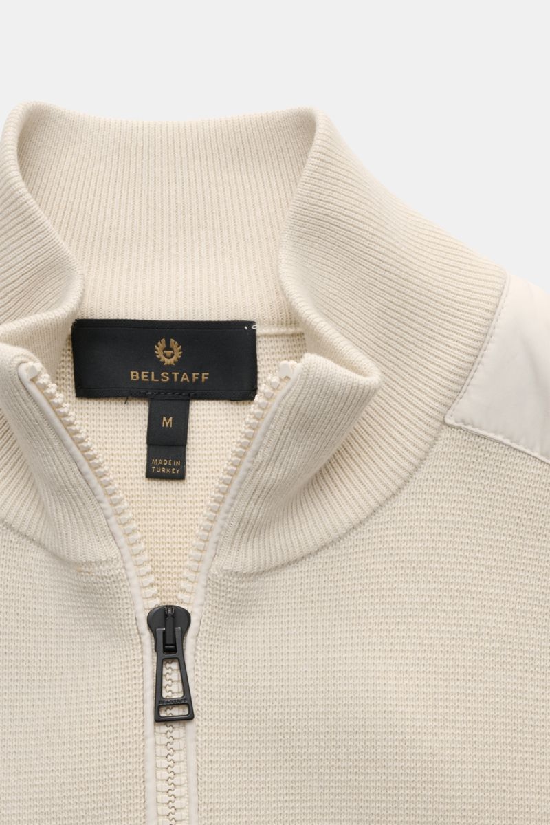Nahaufnahme der Belstaff Strickjacke 'Verge' creme im Slim Fit mit hochschließendem Stehkragen, robustem Reißverschluss, aufgesetzten Schulterpartien aus Kontrastmaterial und BELSTAFF-Weblabel am Oberarm, ideal für Layering-Looks im Frühling aus reiner Ba