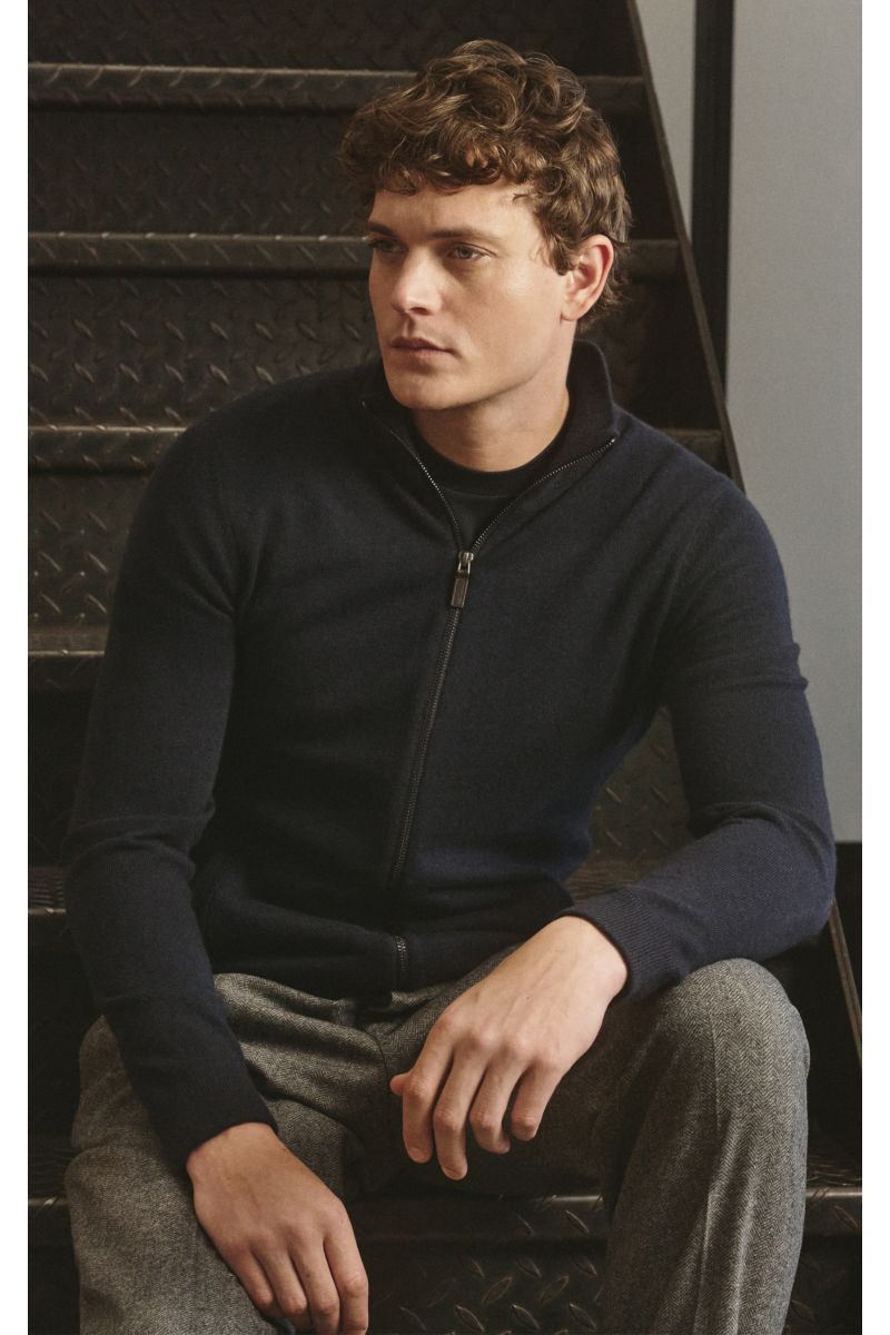 Junger Mann sitzt seitlich auf Treppe, trägt von Braun Cashmere Strickjacke navy mit Stehkragen, Reißverschluss, slim fit, weichem Griff, Leistentaschen, Rippbündchen.
