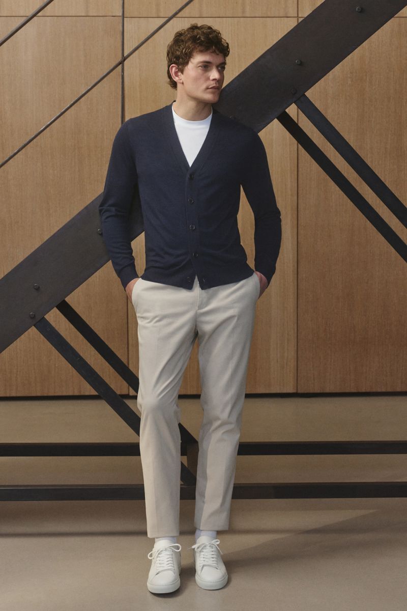 Junger Mann in Frontansicht trägt den von Braun Merino Cardigan navy aus reiner Merinowolle, Regular Fit, mit weichem Griff, feinem Strickbild, Fünf-Knopf-Leiste und elastischen Rippbündchen, kombiniert mit heller Hose und weißen Sneakers.
