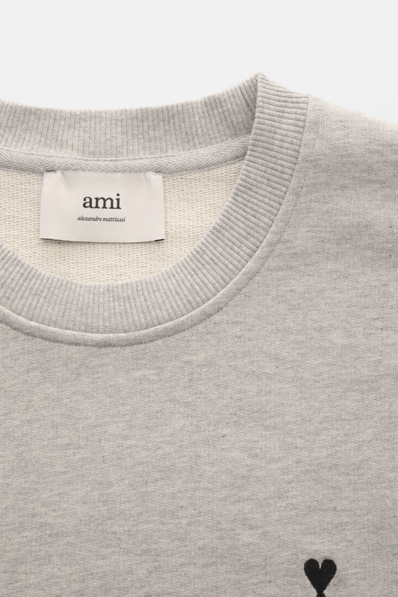 Ami Paris Sweatshirt hellgrau meliert shown from a close-up top-front perspective, featuring a ribbed round neckline, subtle black heart-stitching on the chest, and a visible internal label. Ein dezentes Herz-Stitching auf der Brust verleiht diesem Bau