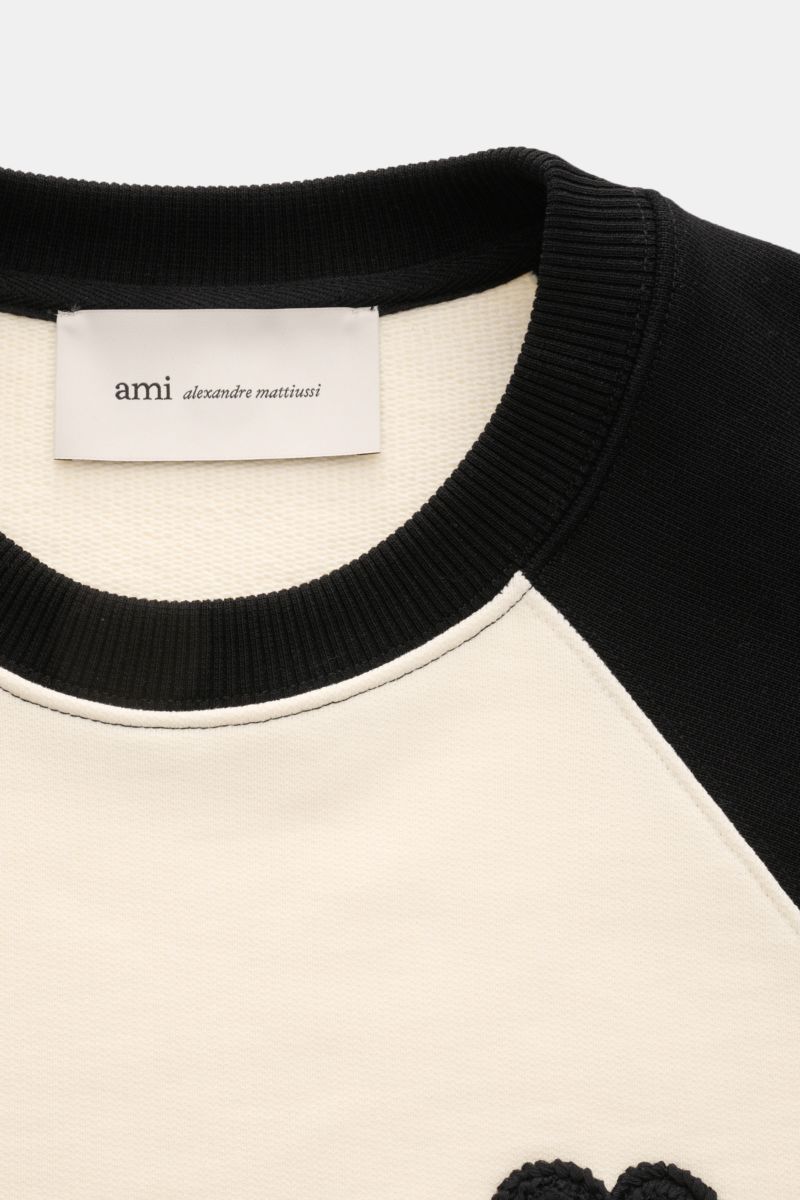 Ami Paris Sweatshirt 'Boxy Fit Crewneck' creme/schwarz shown from a close-up front perspective, highlighting the black ribbed crewneck collar, raglan sleeves in black, and cream body with logo stitching on the chest. Sportive Raffinesse von AMI PARIS: 