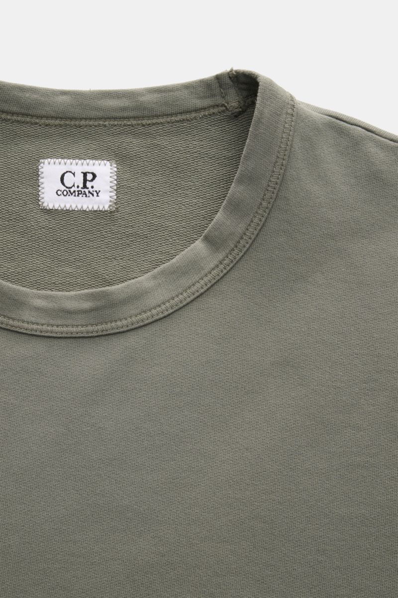 Close-up front top view of a C.P. Company Sweatshirt salbei in a slim fit, garment-dyed light olive green cotton with a round neckline and visible white label, showcasing soft fabric texture and neat stitching. 