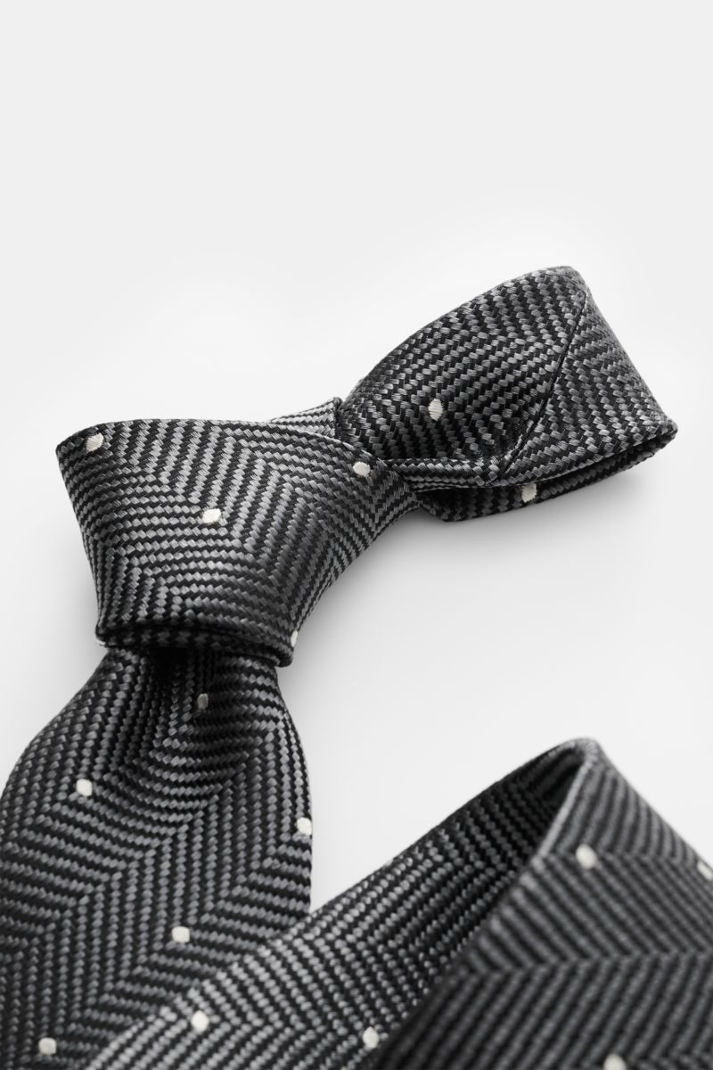 Tom Ford Seidenkrawatte dunkelgrau/schwarz/creme gemustert, in Detailaufnahme schräg von oben, Fischgratmuster mit Punkten, reine Seide, dezenter Glanz, 8 cm breit, made in Italy, Logo-Stitching.