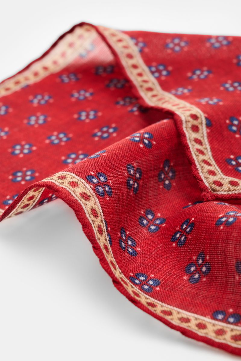 Nahaufnahme aus schräger Perspektive des Rosi Collection Einstecktuchs 'Ted' rot meliert/dunkelblau/beige gemustert aus reiner Wolle mit floralem Print, Borte und rollierten Kanten, Maße ca. 30x30 cm, Made in Italy.