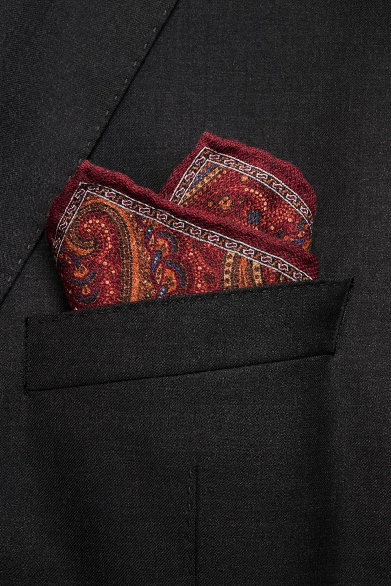 Rosi Collection Seiden-Einstecktuch 'Easy' bordeaux/orange/blau gemustert, reine Seide, Paisley-Muster mit Borte, Medaillon-Rückseite, handrollierte Kanten, soft, 31x31 cm, Made in Italy, steckend in schwarzem Sakko aus leichter Perspektive.