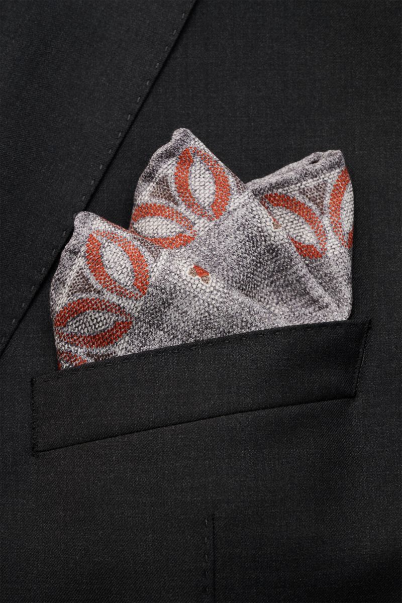 Close-up front view of a Brunello Cucinelli Seiden-Einstecktuch grau/graubraun/rostbraun gemustert tucked into a black suit pocket, showing its floral pattern and texture. reine Seidegeschmeidiger Grifffloral gemustertha