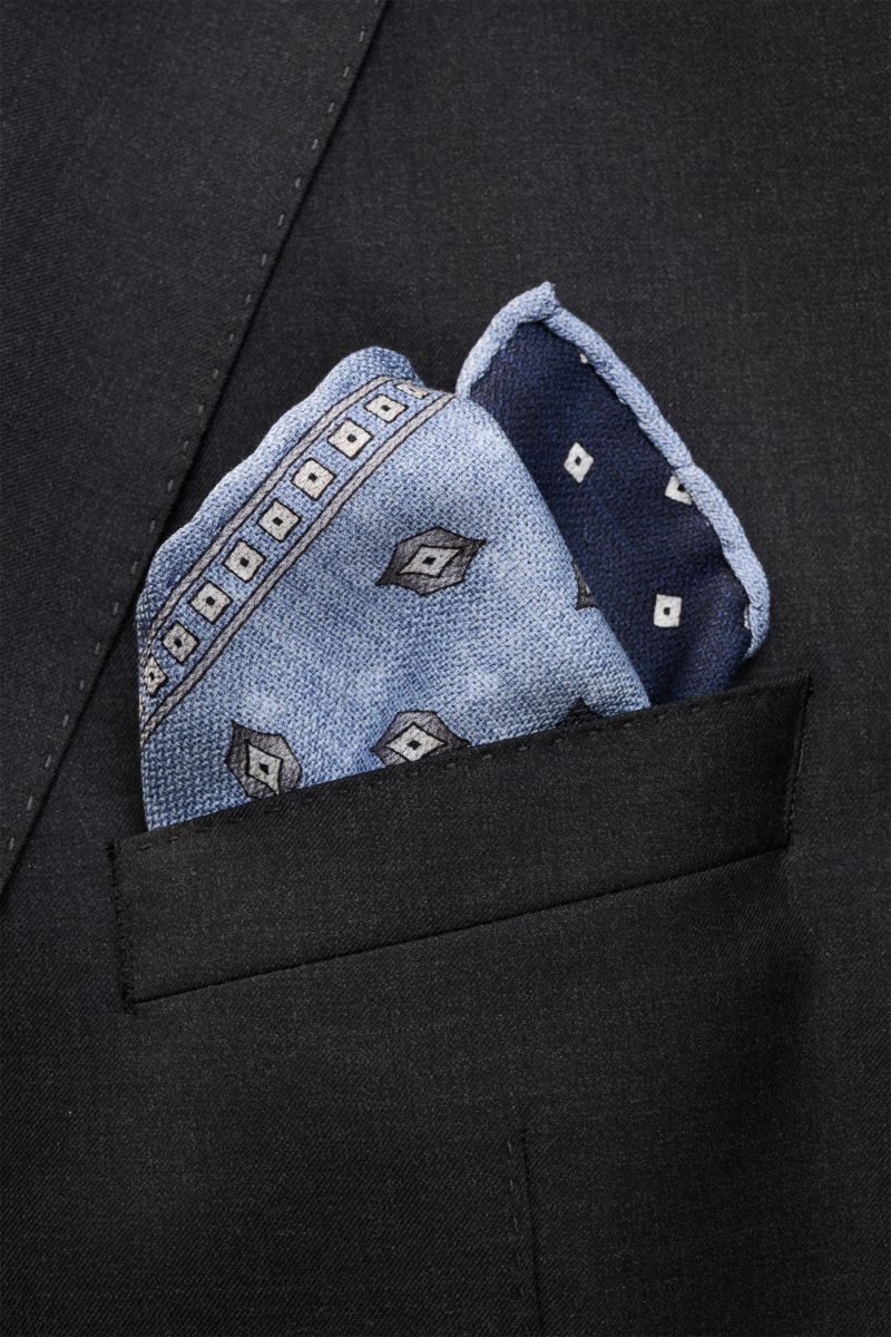 Brustbild aus leichter Draufsicht eines dunkelblauen Sakkos mit rauchblau/blau/grau gemustertem Brunello Cucinelli Seiden-Einstecktuch, reine Seide, geschmeidiger Griff, handrollierte Kanten, Made in Italy, Maße ca. 35x35 cm.