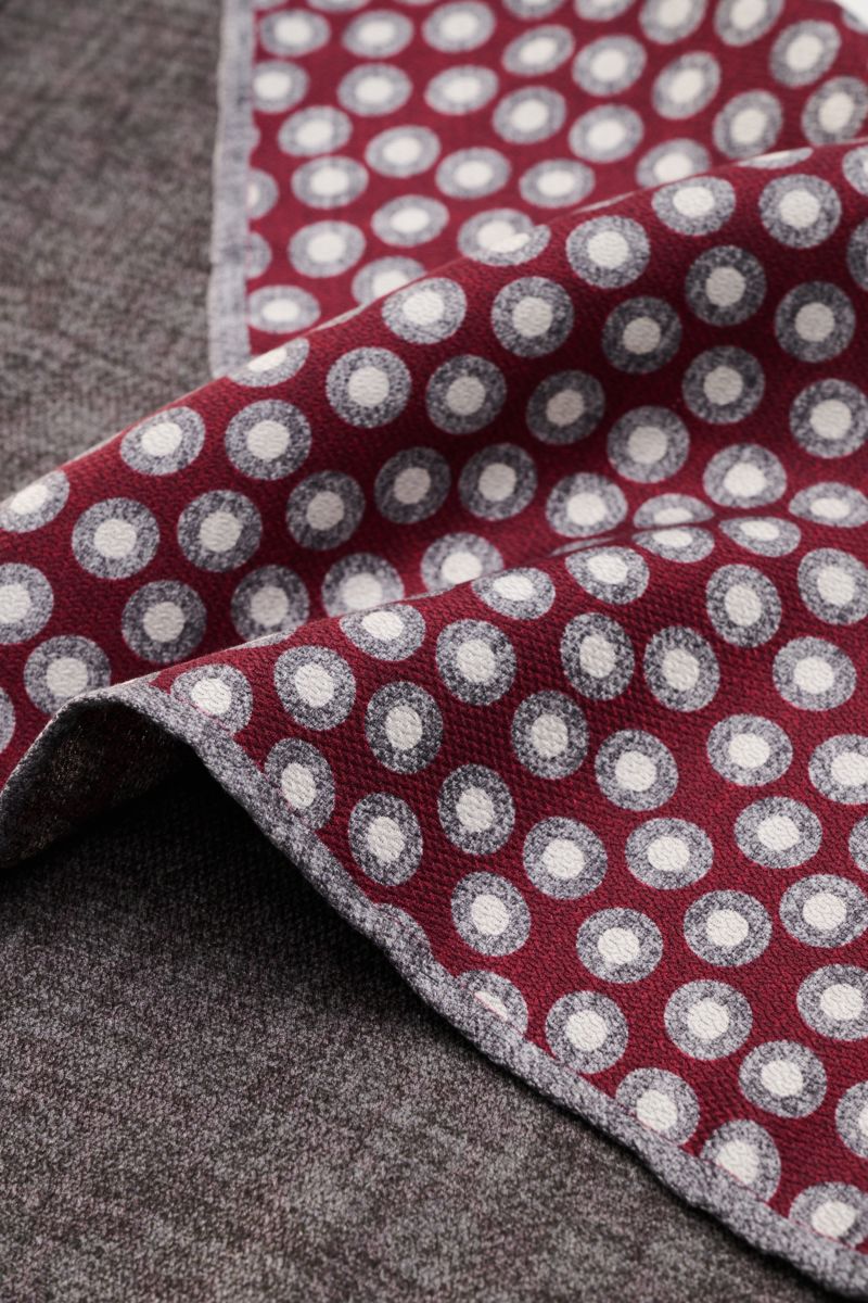 Close-up perspective of the Brunello Cucinelli Seiden-Einstecktuch violett/hellgrau gemustert showing a violet and light gray patterned silk pocket square with a web pattern and hand-rolled edges, placed on a gray fabric surface. reine Seide