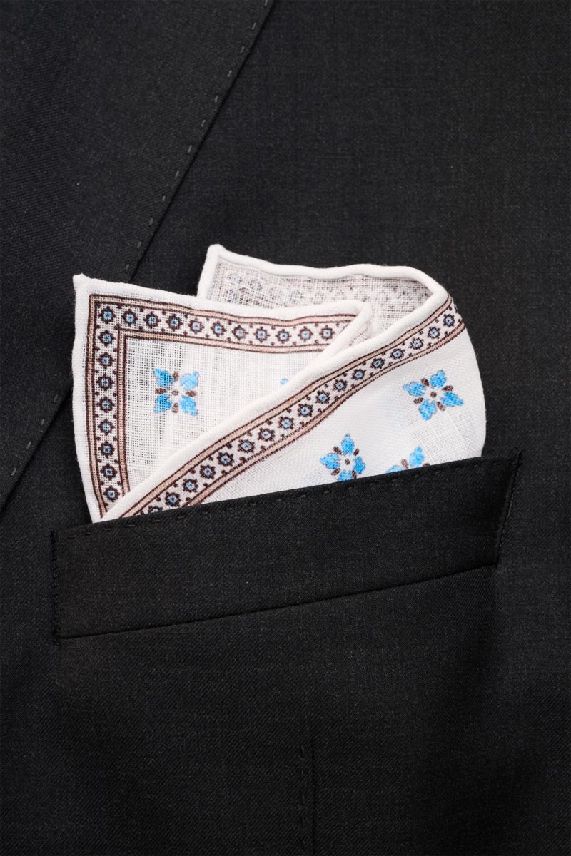 Close-up front view of the Rosi Collection Linen pocket square 'Fox' white/brown/blue patterned tucked into a black suit pocket, featuring pure linen, soft feel, floral print with edging, rolled edges, made in Italy, dimensions 31 cm x 31 cm.