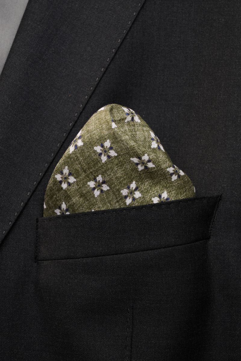 Close-up front view of the Rosi Collection Linen pocket square 'Fox' olive/dark blue/white patterned, pure linen, soft feel, floral print with edging, rolled edges, made in Italy, dimensions 31 cm x 31 cm, tucked neatly into a black suit jacket pocket.
