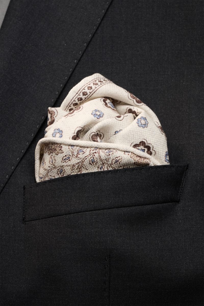Close-up front view of the Rosi Collection Seiden-Einstecktuch 'Easy' beige/dark brown/smoke blue patterned pocket square tucked into a black jacket pocket, featuring pure silk, soft smooth feel, elegant medallion print with border, floral backside, rolle