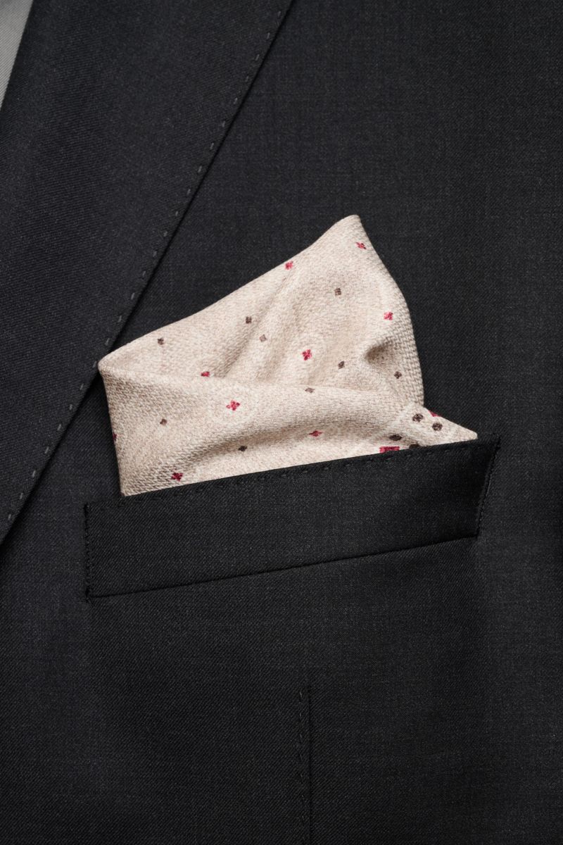 Close-up front view of Brunello Cucinelli Seiden-Einstecktuch beige/weiß/rot gemustert in a black suit pocket, featuring pure silk with graphic print, smooth texture, and hand-rolled edges.

Description: Zeitloser Akzent für besondere Momente: Das Eins