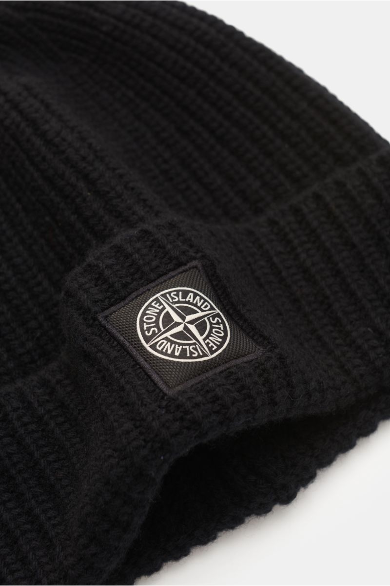 Stone Island Wollmütze schwarz aus reiner Wolle, leicht griffig, moderne Rippstrick-Optik, breiter Umschlag, Front mit STONE ISLAND-Weblabel, Nahaufnahme schräg von oben.