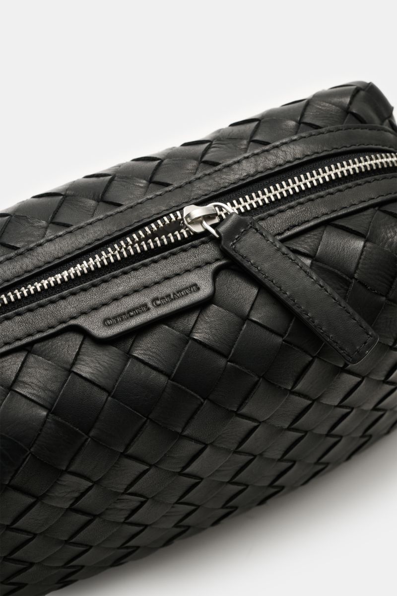 Close-up angled view of the Officine Creative Necessaire schwarz, showing its black woven leather exterior, silver zipper hardware, and embossed logo. Practical travel essential with a spacious main compartment, front zip pocket, and handy wrist strap, si