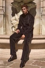 Front view of a male model seated by a stone fountain, dressed in a dark navy corduroy blazer and black cord jogpants from Emporio Armani, paired with black tassel loafers 'Moro Royal' from Henderson. The outfit combines textured fabrics with a sleek, sop