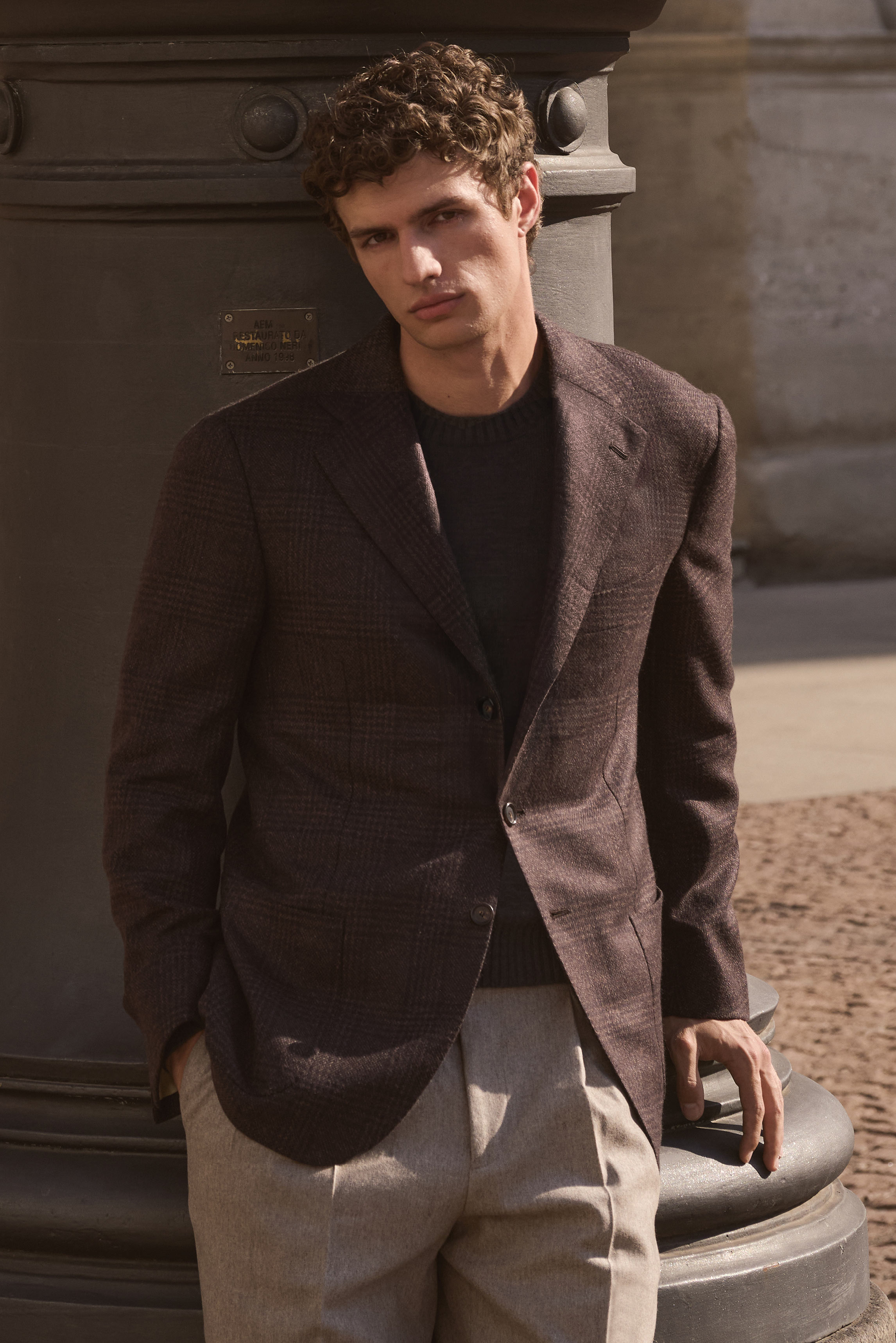 Junger Mann lehnt an Säule, frontal fotografiert, trägt dunkelbraun-schwarz kariertes Cashmere-Sakko von Cesare Attolini, anthrazit melierten Pullover von Fioroni und beige melierte Carrot Fit Wollhose von PT Torino, elegantes Outfit für kühle Tage, Shop 