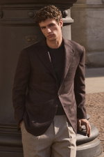 Front view of a model leaning against a column wearing Shop by Look 04 Edition Vol. 19: dark brown and black checkered cashmere blazer from Cesare Attolini, anthracite mottled cashmere crew neck jumper from Fioroni, and beige melange wool 'Carrot Fit' tro