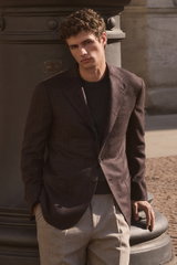 Front view of a model leaning against a column wearing Shop by Look 04 Edition Vol. 19: dark brown and black checkered cashmere blazer from Cesare Attolini, anthracite mottled cashmere crew neck jumper from Fioroni, and beige melange wool 'Carrot Fit' tro