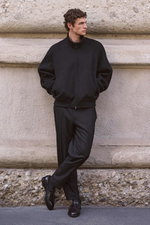 Front view of a model leaning against a stone wall, wearing a black Cashmere Blouson 'Olek' from The Row with high collar, button details, and front zipper, paired with tailored black 'Cava' trousers from Santaniello and black Double Monk 'Opera Flexi' sh