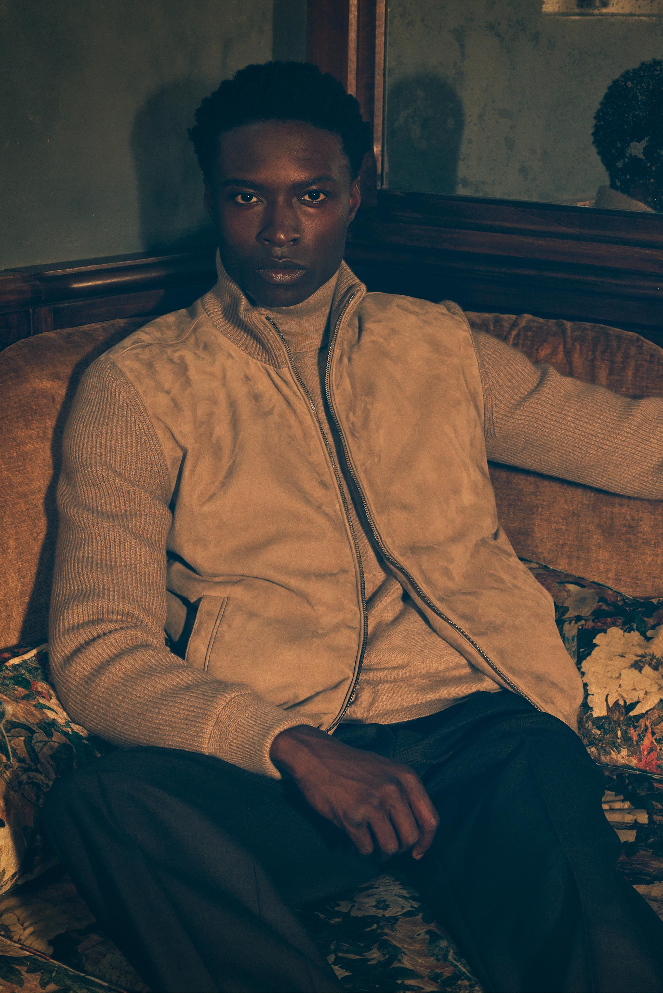 Front view of a model sitting on a vintage sofa wearing taupe cashmere blouson from Agnona over beige cashmere turtleneck sweater from B.F. Knitwear and dark brown melange wool pants from Incotex, showcasing a luxurious, neutral-toned ensemble with soft t