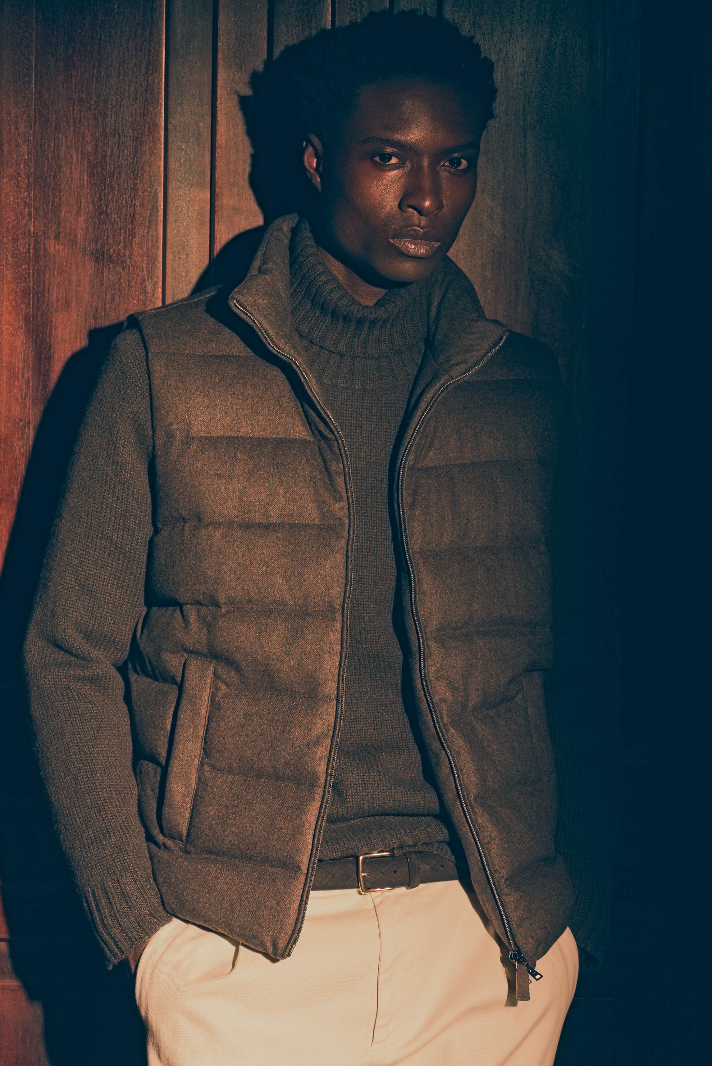 Mann frontal vor Holzwand, Shop by Look 27 Edition Vol. 19 mit braun melierter Herno Daunenweste, dunkelbraunem FTC Cashmere Rollkragenpullover, cremefarbener Dondup Chino 'Ben' und Officine Creative Flechtgürtel, warme Brauntöne und helle Beigetöne, edel