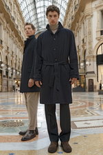 Front view of two male models standing in an ornate indoor gallery, the model in the foreground wearing a navy cashmere coat "Zante" from Stile Latino, belted at the waist, paired with dark pants, and a sand-colored cashmere turtleneck jumper from Bruno M
