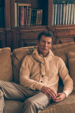 Front view of a seated male model wearing a cream-colored cardigan from Filippo de Laurentiis, cream 'Herrick' chinos from GTA, and a sand-colored bandana from Filippo de Laurentiis around the neck, showcasing a relaxed yet refined look in soft neutral to