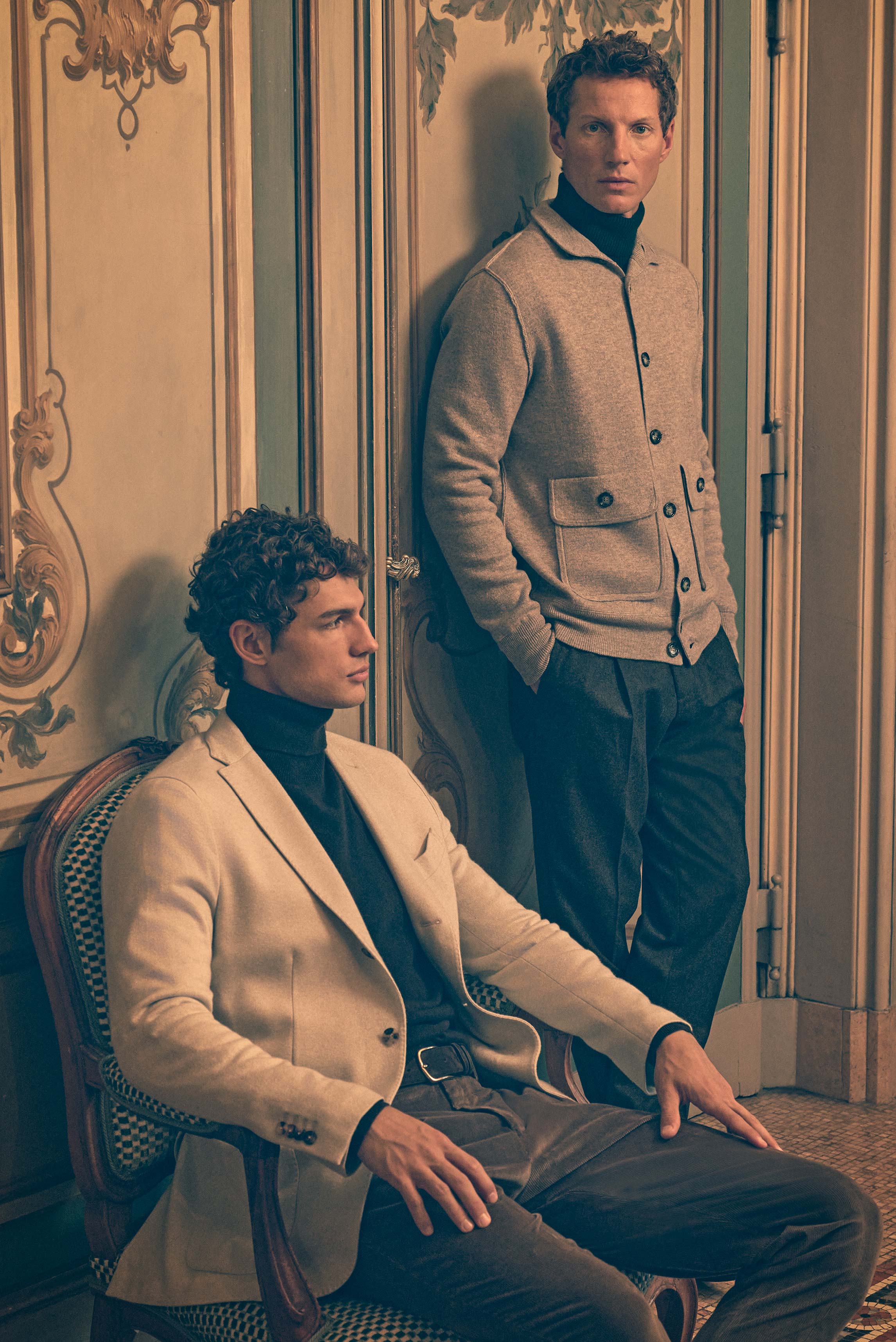 Two men in a vintage room captured from a slight side angle; standing model wears a beige melange knit cardigan from Allude over a dark green cashmere turtleneck by B.F. Knitwear, dark grey melange wool "Carrot Fit" trousers from PT Torino, and a grey-bro